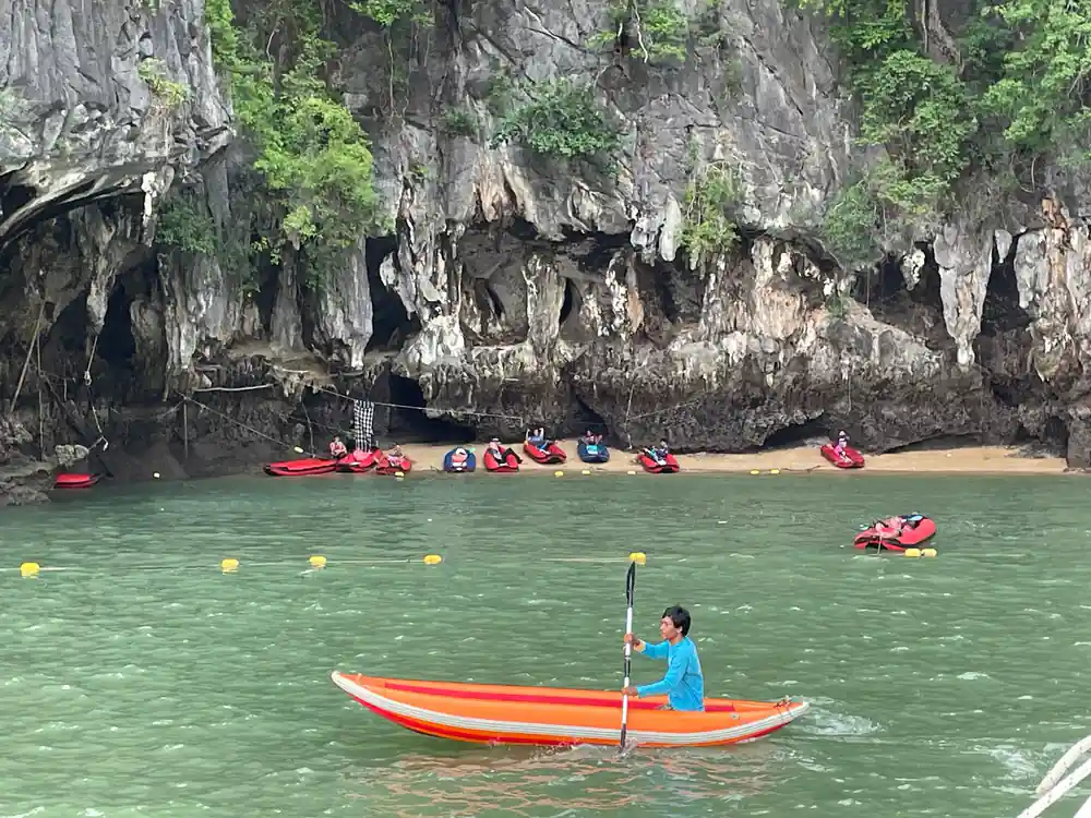 Multiple kayakers and paddleboarders under towering limestone cliff cave